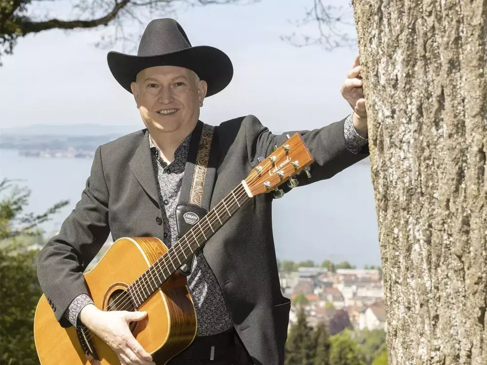 Der Vorarlberger Liedermacher Alfred Bischof lehnt mit schwarzem Cowboyhut und Gitarre an einem Baum vor dem Bodensee und blickt entspannt in die Kamera.